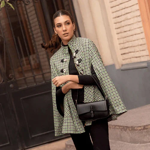 Mujer con capa de cuadros verdes y negros, bolso negro, posando en calle urbana frente a puerta y pared de ladrillos