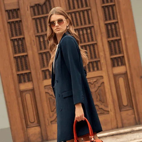 Mujer con abrigo azul marino y gafas de sol, con bolso marrón, frente a puerta de madera detallada