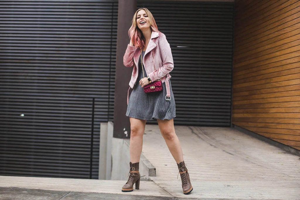 Mujer sonriente con chaqueta rosa, vestido gris y botines marrones en entorno urbano moderno