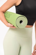 Mujer vestida con top negro y leggings verdes sosteniendo un tapete de yoga enrollado verde