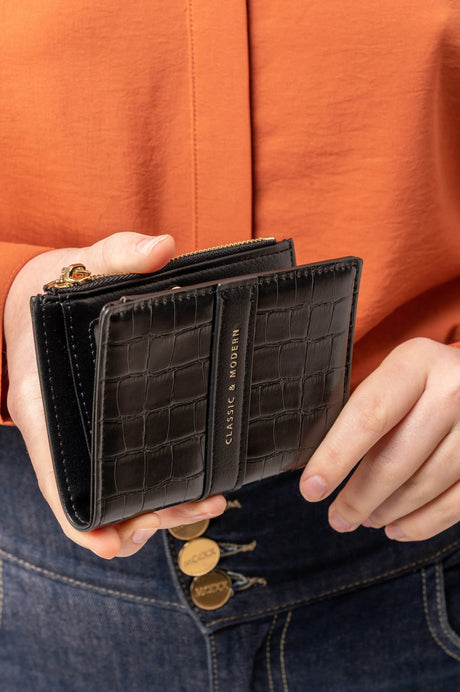 Cartera negra de cuero con diseño de cocodrilo y texto Classic & Modern en manos con camisa naranja y jeans